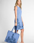 A woman models a light blue sleeveless knit dress with a dark blue botanical leaf pattern, holding a matching blue ribbed tote bag with a dark blue leaf print, set against a light gray studio background.