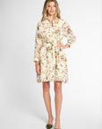 A woman modeling a long-sleeve cream-colored shirt dress with a multicolor floral print and a matching tie belt, paired with olive green pointed-toe flats, against a light gray studio background.