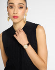 A woman wearing a black sleeveless cable-knit top and a gold-tone wristwatch with a cream dial and brown leather strap, posed against a light gray studio background.