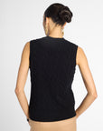 A woman shown from the back wearing a sleeveless black cable-knit sweater vest and beige pants against a light gray studio background.