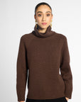 Front view: woman wearing a turtleneck sweater in Chocolate brown with black pants in front of a white background