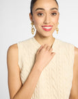 A woman with her hair pulled back wears a cream cable-knit sleeveless top and gold drop earrings against a light gray studio background.