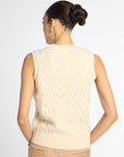 A woman shown from the back wearing a cream cable-knit sleeveless sweater vest and tan pants against a light gray studio background.