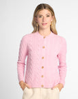 A woman modeling a light pink cable-knit cardigan sweater with gold buttons, paired with cream trousers, against a light gray studio background.