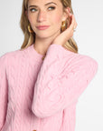 A woman wearing a light pink cable-knit cardigan sweater with gold buttons, shown from the chest up against a plain light gray studio background.
