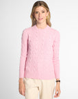 A woman wearing a light pink cable-knit crewneck sweater and cream-colored trousers, standing against a plain light gray studio background.
