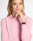 A woman wearing a pink cable-knit mock neck sweater, posing with her hand near her chin against a light gray background.