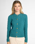 A woman modeling a teal cable-knit cardigan sweater with gold buttons, paired with cream pants, against a light gray studio background.