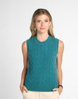 A woman modeling a sleeveless teal cable-knit sweater vest with cream pants, standing against a light gray studio background.