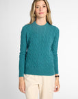 A woman modeling a teal cable-knit crewneck sweater with cream trousers, standing against a light gray studio background.