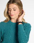A woman wearing a teal cable-knit sweater, gold hoop earrings, and a brown leather watch, posing against a light gray studio background.