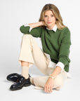 A woman sits cross-legged on a light gray studio floor wearing a green knit sweater over a white collared shirt, cream trousers, beige socks, and navy shoes.