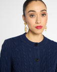 A close-up of a woman wearing a navy blue cable-knit cardigan sweater and gold chain-link drop earrings against a light gray background.