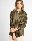 A woman modeling a long-sleeve button-up shirt with vertical olive green and beige stripes, paired with cream pants, against a light gray studio background.