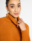 A close-up of a woman wearing a chunky knit turtleneck sweater in a warm camel orange color, paired with chunky gold chain drop earrings, against a plain light gray background.