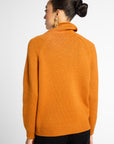 A woman shown from the back wearing a bright orange knit sweater with a turtleneck and raglan sleeves, paired with black pants, against a light gray studio background.