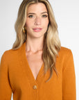 A woman with blonde wavy hair wearing a mustard orange knit cardigan sweater with brown buttons, photographed against a light gray studio background.