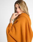 A woman with blonde hair wearing a warm orange knit poncho wrap, photographed against a clean light gray studio background.