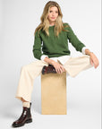 A woman models a green crewneck sweater, cream wide-leg trousers, and dark brown leather ankle boots while sitting on a light wood block against a white studio background.