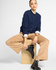 A model sits on a light wood block wearing a navy blue cable-knit cardigan over a white collared shirt, beige wide-leg trousers, and black Chelsea boots, against a light gray studio background.