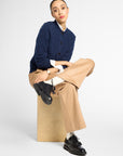A model sits on a light brown cardboard box wearing a navy blue cable-knit cardigan over a white collared shirt, paired with beige wide-leg trousers and black leather shoes, against a plain white studio background.
