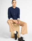 A model sits on a cardboard box wearing a navy blue cable-knit cardigan over a white collared shirt, beige wide-leg trousers, white socks, and black leather shoes against a light gray studio background.