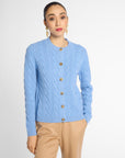 A woman modeling a light blue cable-knit cardigan sweater with brown buttons, paired with beige trousers, against a plain light gray studio background.