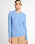 A woman modeling a light blue cable-knit crewneck sweater with beige trousers, standing against a plain light gray studio background.