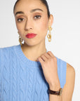 A close-up of a woman wearing gold drop earrings with textured, organic loops, paired with a light blue cable-knit sweater vest and a brown leather watch.