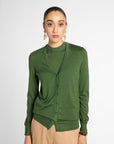 A woman modeling a green long-sleeve button-front cardigan over a matching green top, paired with beige trousers, against a light gray studio background.