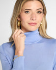 A woman with blonde hair wearing a light blue turtleneck sweater and large gold hoop earrings, posing against a light gray background.