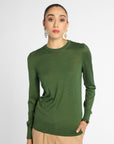 A woman wearing a fitted long-sleeve crewneck sweater in olive green, paired with beige trousers, against a plain light gray studio background.