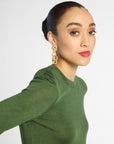 A woman wearing a green long-sleeve knit top and large gold statement earrings, photographed against a light gray studio background.