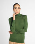 A woman wearing a fitted ribbed green turtleneck sweater and beige trousers, standing against a light gray studio background.
