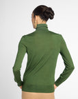 A woman shown from the back wearing a fitted green turtleneck sweater and beige pants against a light gray studio background.