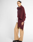 Side view: A woman wearing a chunky knit burgundy shawl draped over her shoulders, styled over a white top, against a plain light gray studio background