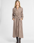 A woman modeling a long brown and beige houndstooth belted coat dress with pockets, paired with dark brown boots, against a light gray studio background.