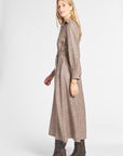 A woman modeling a long-sleeved, collared plaid midi dress in brown and beige tones, paired with dark brown heeled boots, against a light gray studio background.