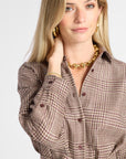 A woman wearing a brown and beige plaid button-up shirt with a tie waist, styled with gold jewelry against a light gray background.