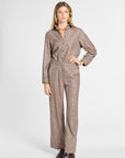 A woman modeling a brown and beige plaid long-sleeve button-up shirt and matching wide-leg trousers, styled with brown leather shoes and gold jewelry against a light gray studio background.