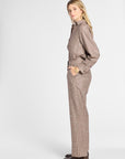 A woman modeling a matching two-piece plaid outfit in a side profile against a light gray studio background. The set consists of a long-sleeve collared shirt and matching wide-leg trousers in a brown and beige check pattern, paired with dark brown leather shoes.