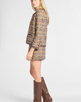 A woman modeling a tweed jacket and matching mini skirt in a multicolor woven pattern, paired with brown knee-high leather boots, against a light gray studio background.