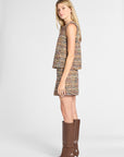 A woman modeling a sleeveless tweed mini dress with a matching mini skirt in a multicolor bouclé pattern, paired with brown knee-high leather boots, against a light gray studio background.