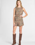 A woman modeling a sleeveless tweed romper with matching shorts in a multicolor woven pattern, paired with brown knee-high leather boots, against a light gray studio background.