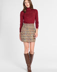 A woman modeling a burgundy turtleneck long-sleeve top and a multicolor tweed mini skirt, paired with brown knee-high leather boots, against a light gray studio background.