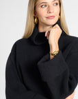 A woman wearing a black ribbed knit mock neck sweater, styled with gold hoop earrings, a gold watch, and a gold bracelet, against a light gray studio background.