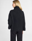 A woman shown from the back wearing a black knit turtleneck sweater and black wide-leg trousers against a light gray studio background.