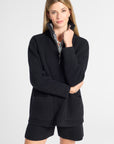 A woman modeling a black knit quarter-zip sweater with a striped inner collar and matching black shorts against a light gray studio background.