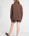 A woman shown from the back wearing a brown knit quarter zip sweater and matching brown shorts, paired with brown shoes, against a light gray studio background.