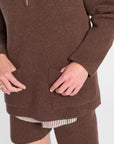 A close-up of a person wearing a brown ribbed knit sweater dress with a quarter-zip neckline and front patch pockets, paired with striped shorts against a plain light background.